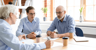 A British engineering consultancy founder and his adviser sit across from acquirer representatives in a small meeting room inside an older commercial building, mid-negotiation over deal synergies, afternoon light filtering through a sash window.