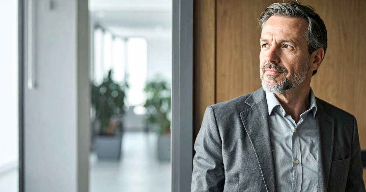 A founder in their mid-50s stands quietly at an office doorway in late-afternoon light, considering the next stage of their business.