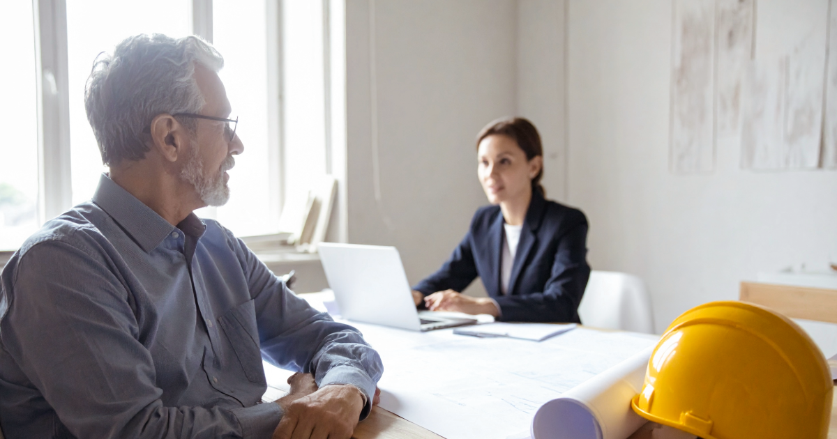 A consultancy founder in his fifties stepping back as a colleague leads a client meeting in a daylit office.