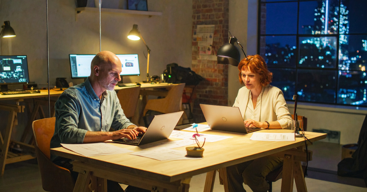 Two SaaS co-founders working late at a desk lit by a desk lamp, London skyline visible through a Victorian sash window behind them.