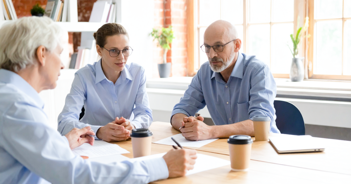 A British engineering consultancy founder and his adviser sit across from acquirer representatives in a small meeting room inside an older commercial building, mid-negotiation over deal synergies, afternoon light filtering through a sash window.