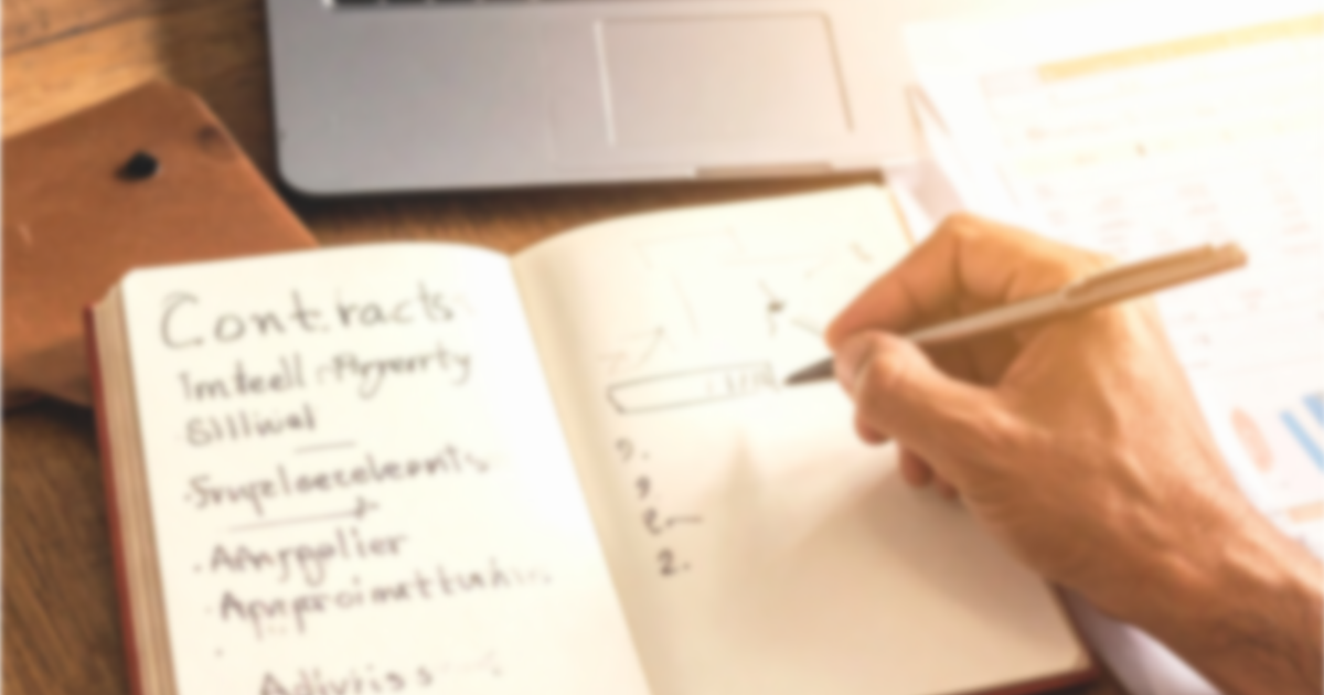 A founder making handwritten notes in an open notebook at a desk, with a laptop showing a spreadsheet open in the background