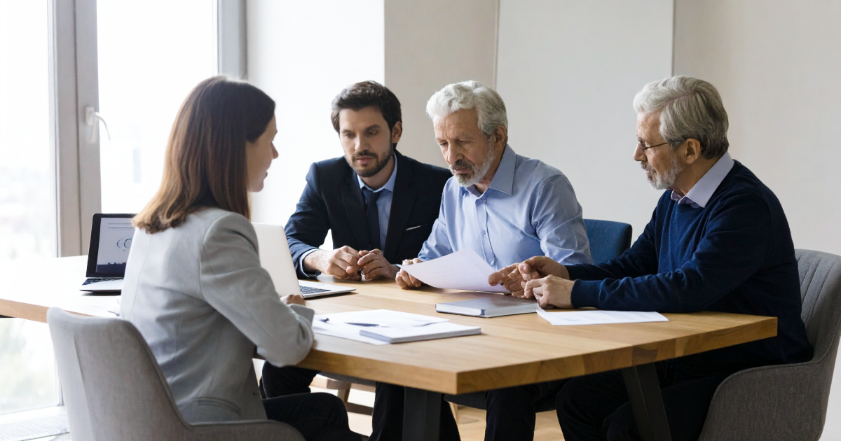 Founder and shareholder reviewing acquisition research list across a boardroom table — daylight interior, documents and laptop open.